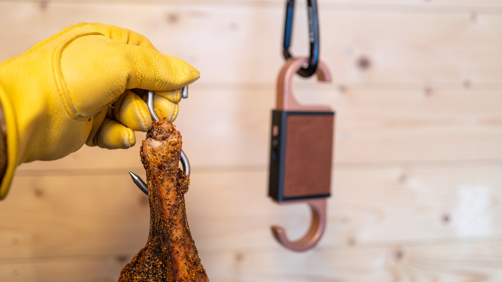Hand wearing a yellow glove holding a parma ham with a hanging meat scale in the background.