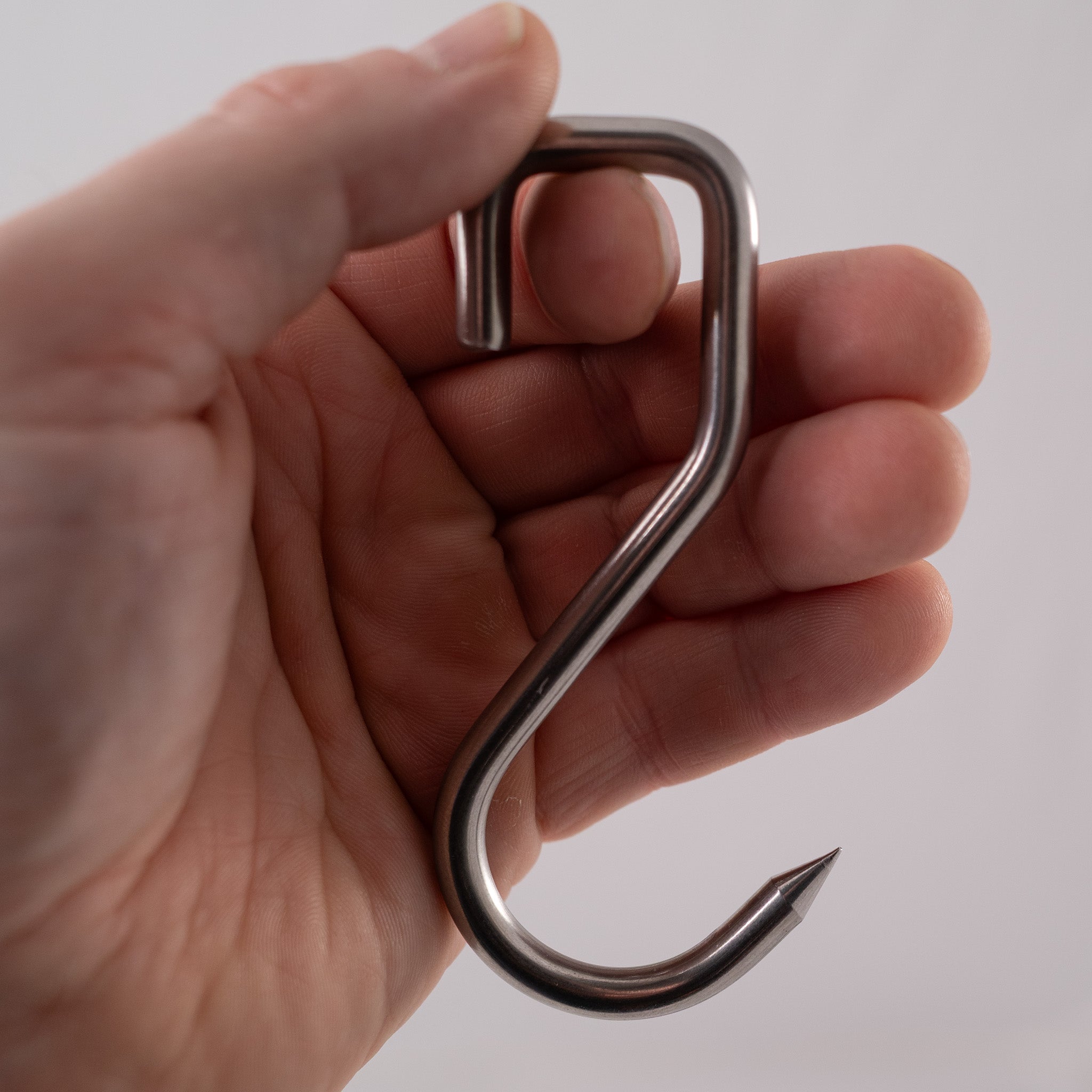 Hand holding a metallic s-hook, a food safe sharp stainless steel meat hook on a white background