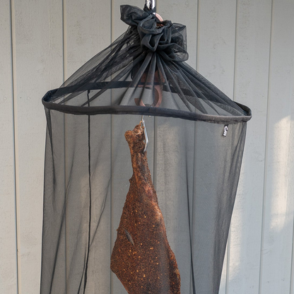 Parma ham hanging inside a black mesh protective cover against a wooden wall.