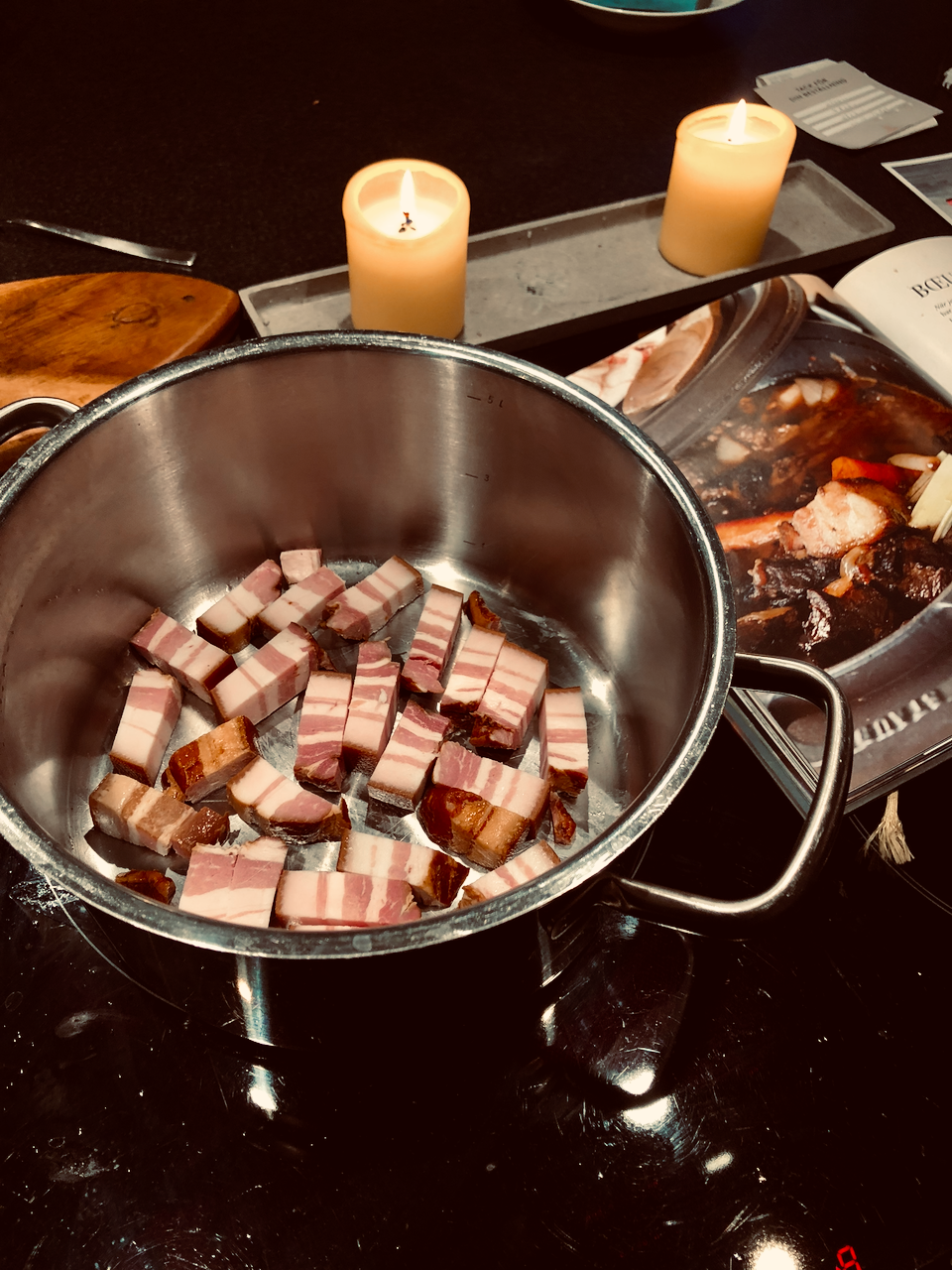 Metal pot with chopped smoked pork belly meat on a dark surface with candles in the background, boeuf bourguignon