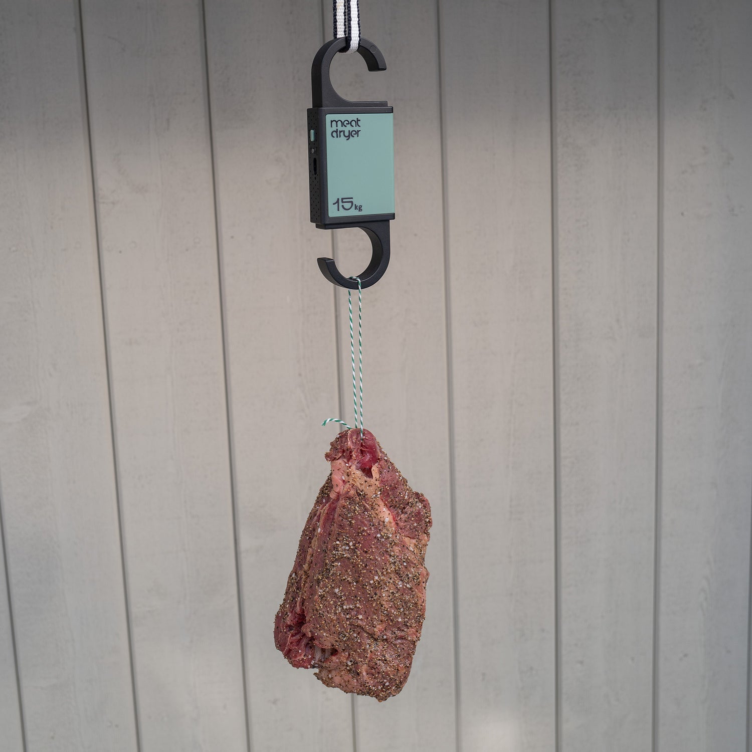 Biltong beef meat hanging from a green Meatdryer hook with a weight , temperature and humidity indicator against a grey background